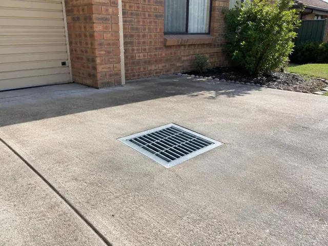 Dry concrete driveway with a clean metal drain grate near a brick house in Kingswood